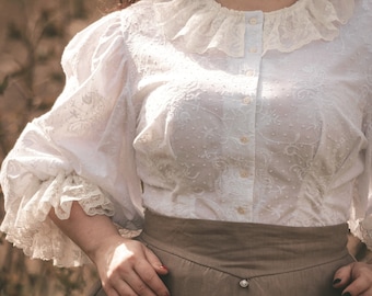 May include: A white blouse with a lace collar and embroidered details. The blouse has a button-up front and long sleeves with lace trim. The blouse is paired with a brown skirt.