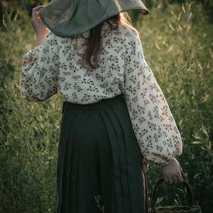May include: A person wearing a green wide-brimmed hat and a white blouse with a floral pattern, standing in a field of tall green grass. They are holding a woven basket.