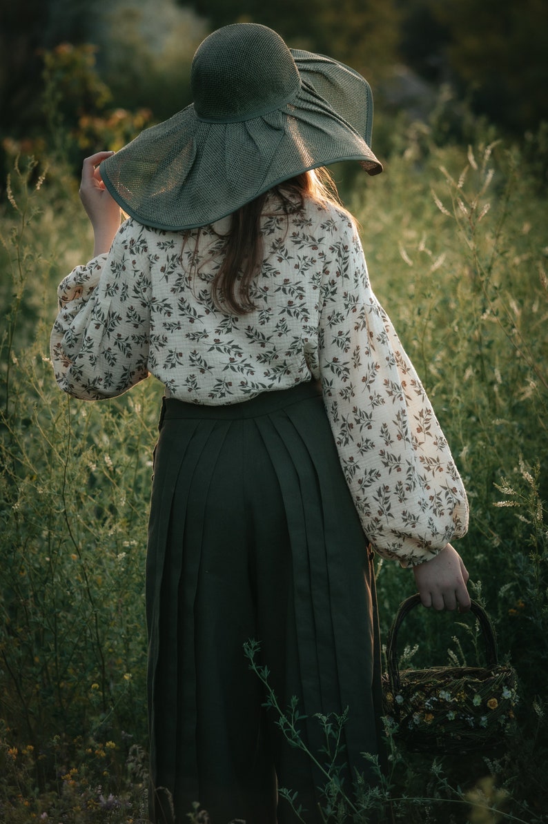May include: A person wearing a green wide-brimmed hat and a white blouse with a floral pattern, standing in a field of tall green grass. They are holding a woven basket.