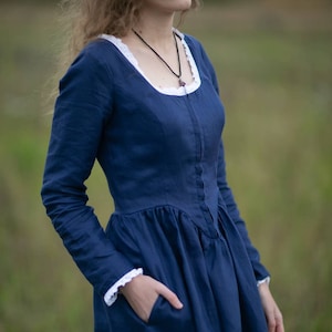 May include: A woman wearing a long-sleeved, navy blue linen dress with a white lace trim around the neckline and cuffs. The dress has a fitted bodice and a full skirt. She is wearing a black necklace with a small pendant.