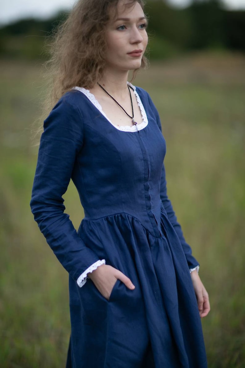 May include: A woman wearing a long-sleeved, navy blue linen dress with a white lace trim around the neckline and cuffs. The dress has a fitted bodice and a full skirt. She is wearing a black necklace with a small pendant.