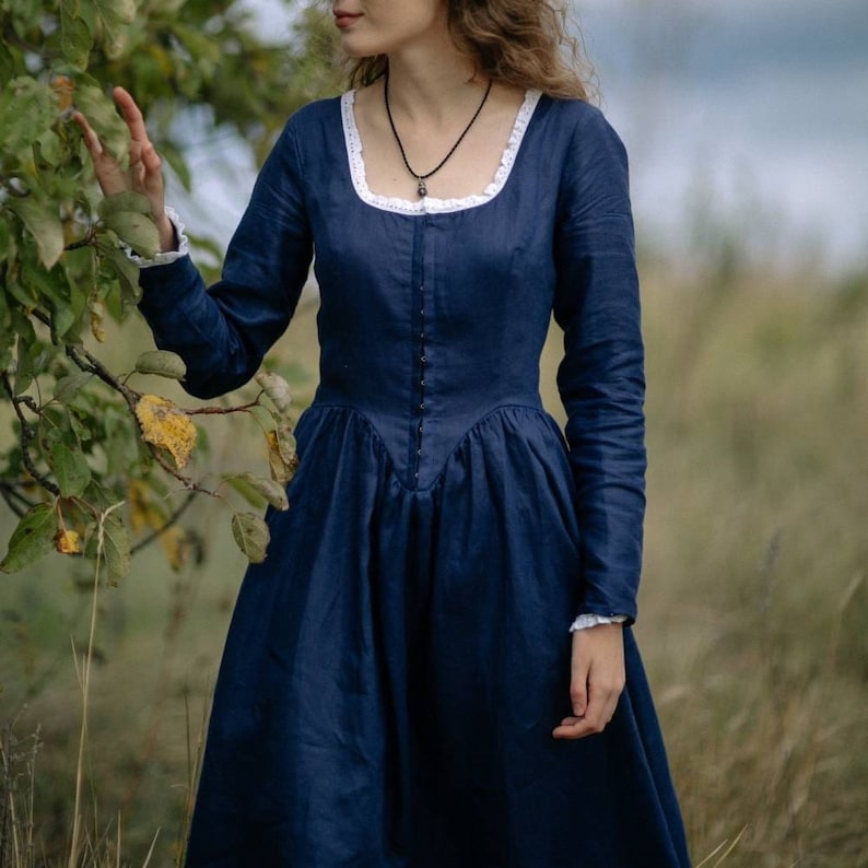 May include: A woman wearing a long-sleeved, dark blue dress with a white lace neckline. The dress has a fitted bodice and a full skirt. She is standing in a field of long grass.