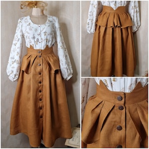 Può includere: Una gonna marrone con una blusa bianca con stampa floreale. La gonna ha un'alta vita e uno strato di volant in alto. La gonna è abbottonata sul davanti.