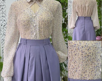 May include: A long-sleeved blouse with a cream and blue floral pattern, featuring a pointed collar, a triangular cutout at the neckline, and a button-down front, paired with a solid lavender high-waisted skirt with vertical pleats.