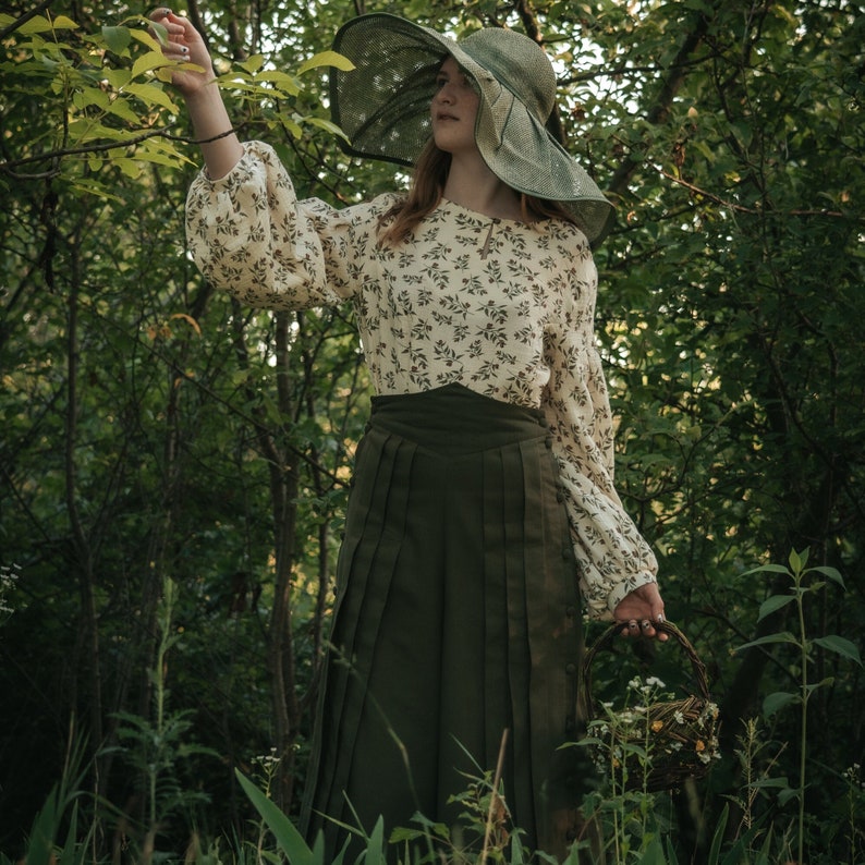 May include: A woman wearing a white floral blouse and a green skirt with a wide-brimmed straw hat, standing in a forest setting. She is holding a basket of flowers.