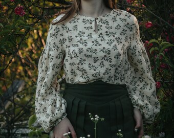 May include: A woman wearing a white blouse with a floral pattern and a dark green skirt stands in a garden with pink flowers.