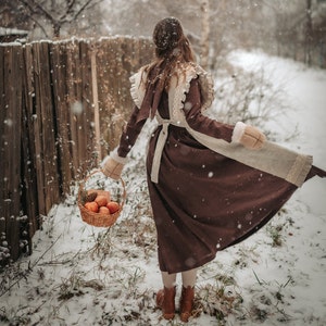 May include: A woman in a brown dress with a white lace overlay and a white fur trim walks through a snowy forest carrying a basket of apples.