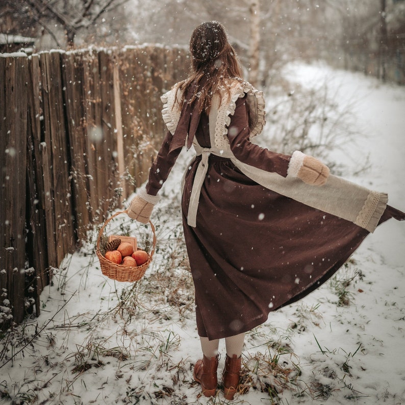 May include: A woman in a brown dress with a white lace overlay and a white fur trim walks through a snowy forest carrying a basket of apples.