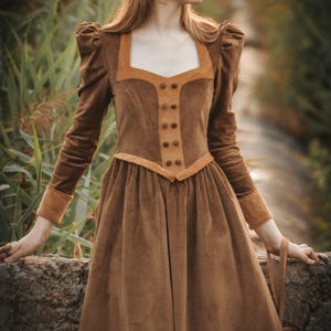 Puede incluir: Un vestido de terciopelo marrón con un corpiño ajustado y una falda completa. El vestido tiene un escote cuadrado y mangas largas con hombros abullonados. El corpiño está acentuado con un panel de ante marrón y botones.