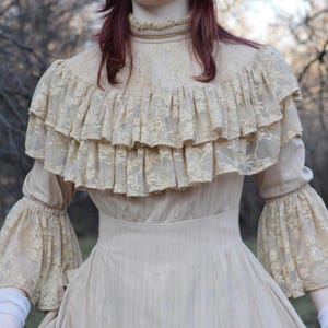 May include: A cream-colored vintage dress with a high ruffled collar and tiered lace detailing. The dress features long sleeves with flared lace cuffs and a fitted bodice. The skirt has vertical stripes, creating a classic, elegant silhouette.
