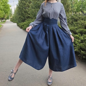 Può includere: Una camicetta grigia a maniche lunghe con scollatura a goccia e culotte blu navy a vita alta e gamba larga. La culotte ha pieghe in vita. L'outfit è abbinato a scarpe Mary Jane grigie.