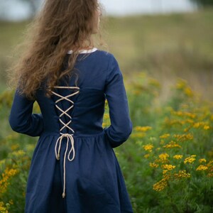 May include: A woman wearing a navy blue dress with a lace-up back stands in a field of yellow wildflowers.