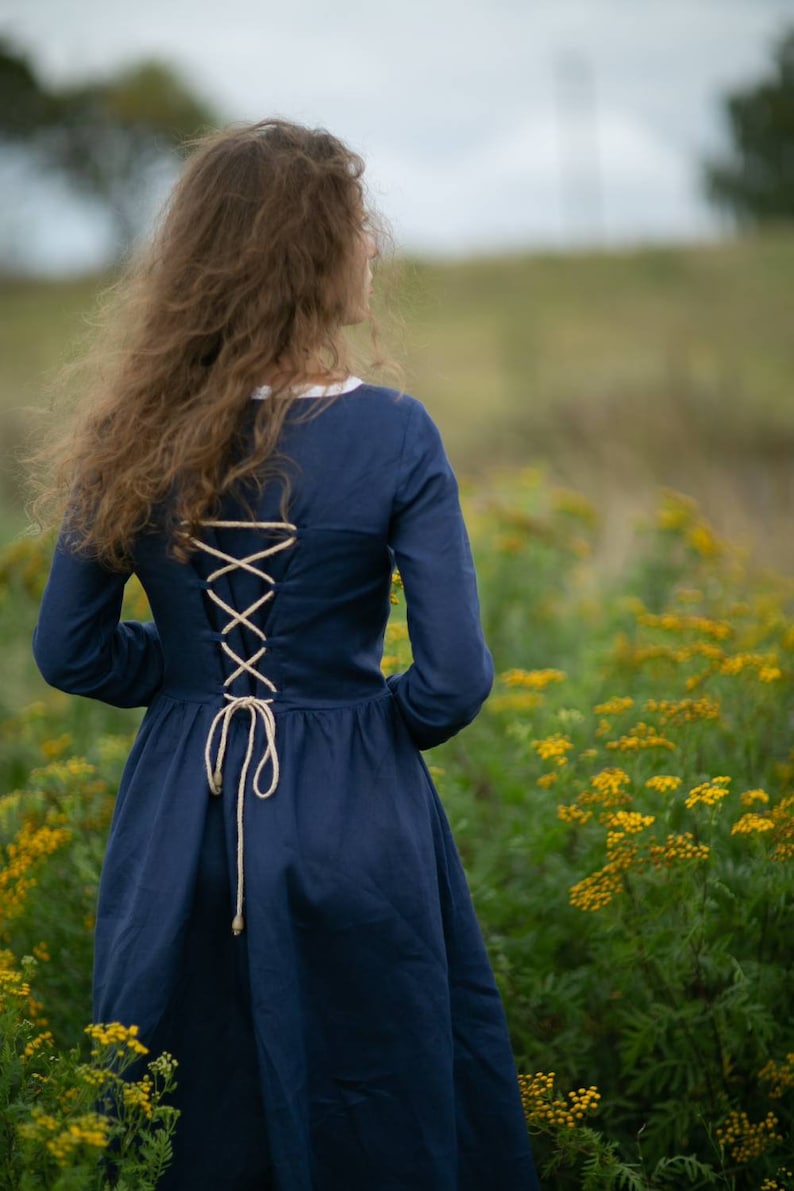 May include: A woman wearing a navy blue dress with a lace-up back stands in a field of yellow wildflowers.