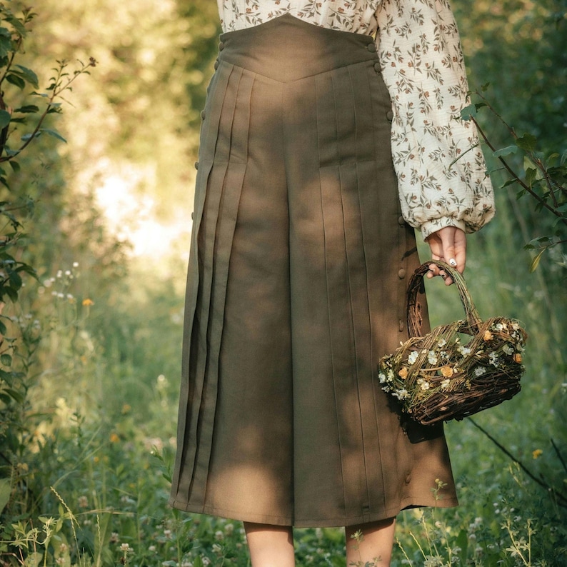 May include: A woman wearing a white blouse with a floral pattern and a pair of dark green, pleated culottes. She is holding a wicker basket filled with dried flowers.