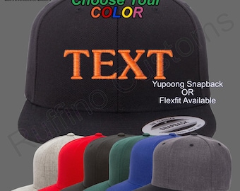 May include: A black baseball cap with the word "TEXT" embroidered in orange. The cap is available in a variety of colors, including red, grey, green, blue, and black. The cap is made by Yupoong and is available in a Flexfit style.