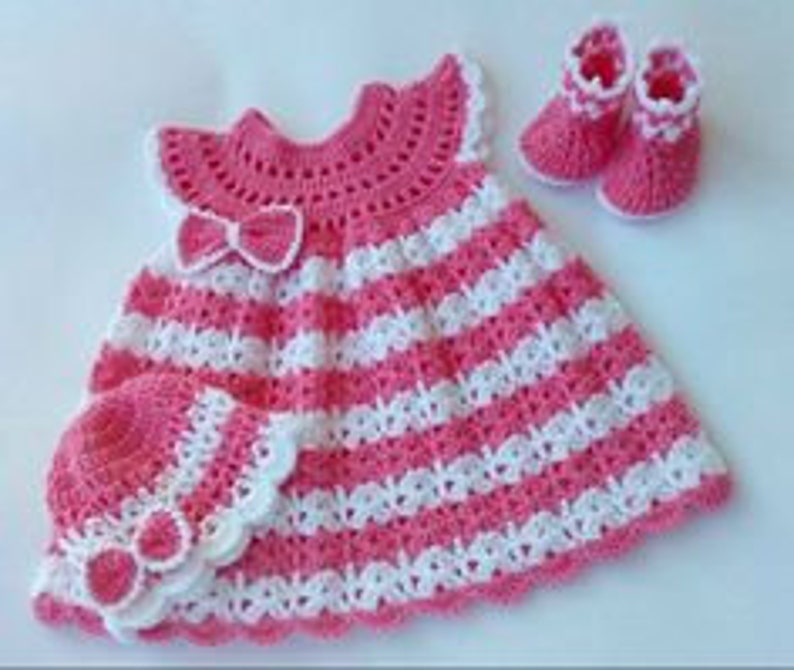 K&ouml;nnte beinhalten: Ein gestricktes Babykleid in rosa und wei&szlig; mit passendem Hut und Schuhen. Das Kleid hat einen R&uuml;schenkragen und eine Schleife.