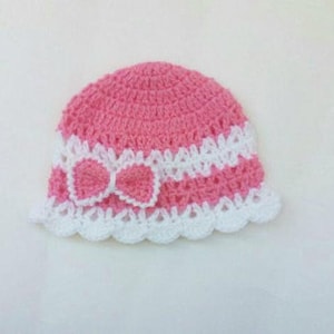 K&ouml;nnte beinhalten: Eine rosa und wei&szlig;e geh&auml;kelte Baby-M&uuml;tze mit einer wei&szlig;en Schleife.