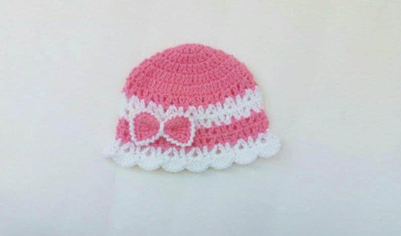 K&ouml;nnte beinhalten: Eine rosa und wei&szlig;e geh&auml;kelte Baby-M&uuml;tze mit einer wei&szlig;en Schleife.