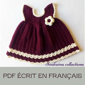 May include: A burgundy crochet baby dress with a white crocheted flower and white trim. The dress is sleeveless and has a round neckline. The text "Soukaïna collections PDF ÉCRIT EN FRANÇAIS" is at the bottom of the image.