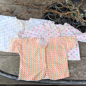 May include: Three white baby shirts with different coloured polka dots. The shirts have a button-up front and short sleeves. The polka dots are blue, pink, and orange and green.