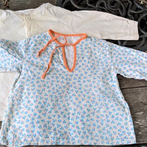 May include: Two vintage baby shirts. One is white with a button closure. The other is white with a blue floral pattern and an orange trim.