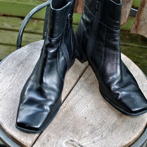 May include: A pair of black leather ankle boots with a square toe and zipper closure. The boots are sitting on a wooden surface.