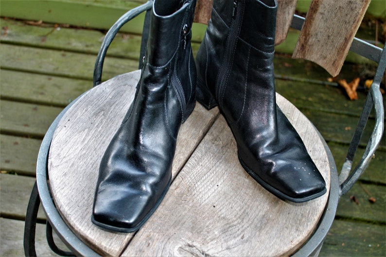 May include: A pair of black leather ankle boots with a square toe and zipper closure. The boots are sitting on a wooden surface.