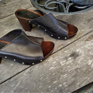 May include: A pair of dark brown leather heeled clogs with a wooden platform and silver studs. The clogs are on a wooden surface.