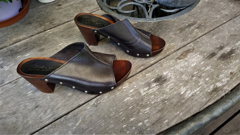 May include: A pair of dark brown leather heeled clogs with a wooden platform and silver studs. The clogs are on a wooden surface.