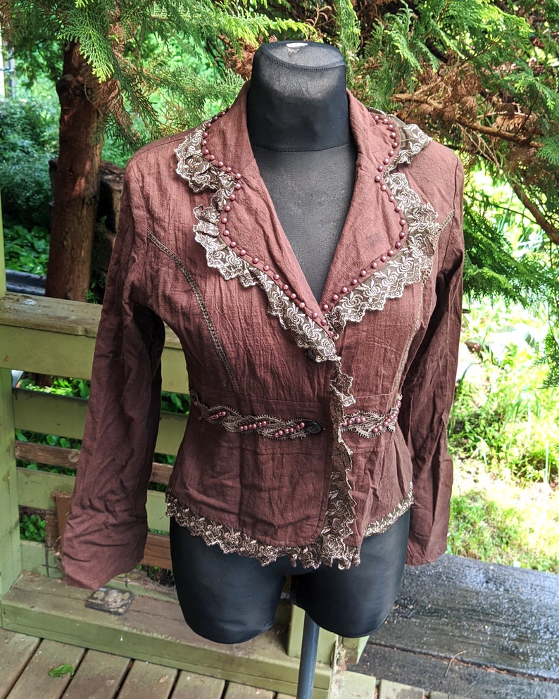 May include: A brown jacket with a lace trim and beaded accents. The jacket has a button closure and long sleeves.