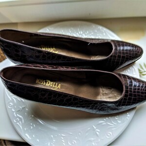 May include: A pair of brown crocodile-embossed leather shoes with a pointed toe. The shoes are on a white plate with a floral design. The brand name "Miss Della" is visible on the insole of one shoe.