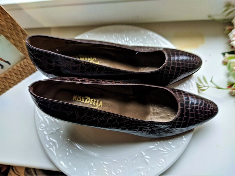 May include: A pair of brown crocodile-embossed leather shoes with a pointed toe. The shoes are on a white plate with a floral design. The brand name "Miss Della" is visible on the insole of one shoe.