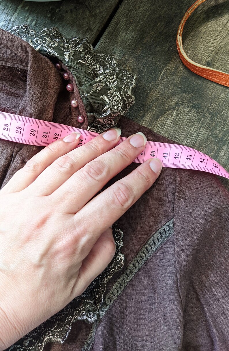 May include: A brown linen top with a green lace trim and pink beads. A pink measuring tape is placed across the top, showing measurements from 28 to 45 inches.