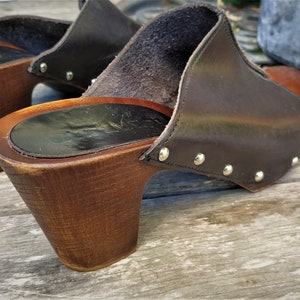 May include: A pair of brown leather clogs with wooden heels. The clogs have a pointed toe and are decorated with silver studs.