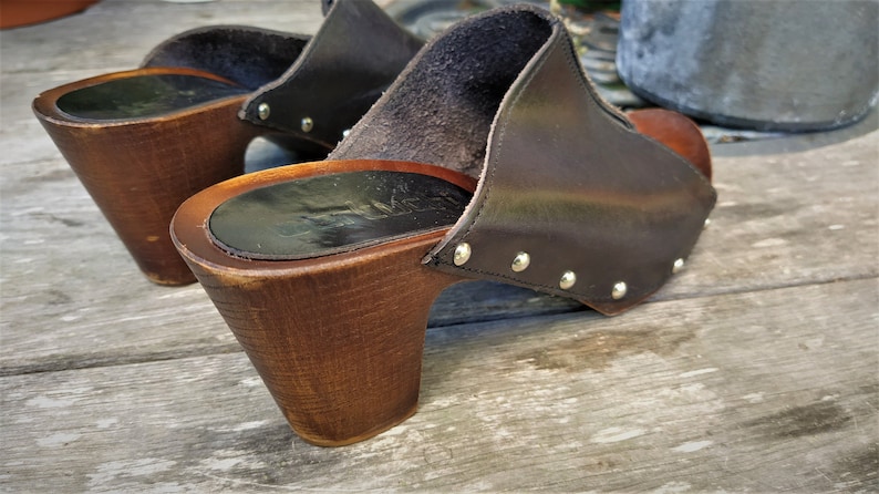 May include: A pair of brown leather clogs with wooden heels. The clogs have a pointed toe and are decorated with silver studs.