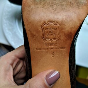 May include: Close-up of the bottom of a brown leather shoe with a black embossed pattern. The sole is labeled "Calcado de Couro" and "Lunik" with the text "Fabricado em Portugal" and the size "6".