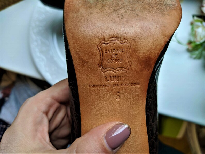 May include: Close-up of the bottom of a brown leather shoe with a black embossed pattern. The sole is labeled "Calcado de Couro" and "Lunik" with the text "Fabricado em Portugal" and the size "6".