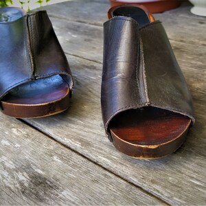 May include: A pair of brown leather clogs with wooden soles. The clogs have a peep-toe design and are sitting on a wooden surface.