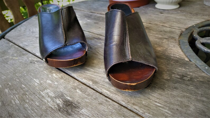 May include: A pair of brown leather clogs with wooden soles. The clogs have a peep-toe design and are sitting on a wooden surface.