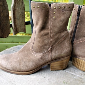 May include: A pair of brown suede ankle boots with a zipper on the side and a wooden heel. The boots have decorative studs on the top of the shaft and the brand name "Tamaris" is visible on the side.