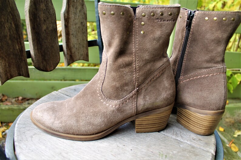 May include: A pair of brown suede ankle boots with a zipper on the side and a wooden heel. The boots have decorative studs on the top of the shaft and the brand name "Tamaris" is visible on the side.