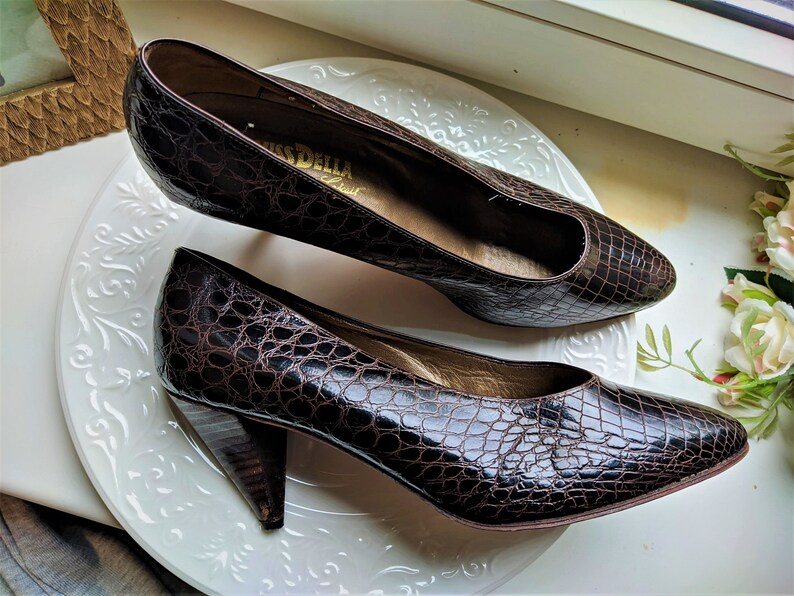 May include: A pair of brown crocodile-embossed leather pumps with a pointed toe and a low block heel. The shoes are on a white plate and the brand name "Miss Della" is visible on the insole.