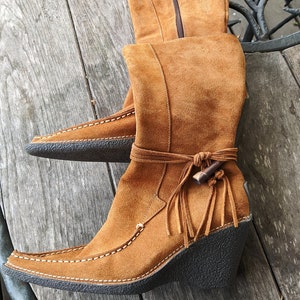 May include: A pair of brown suede boots with a fringe detail and a wedge heel. The boots have a zipper on the side and a white stitching detail.