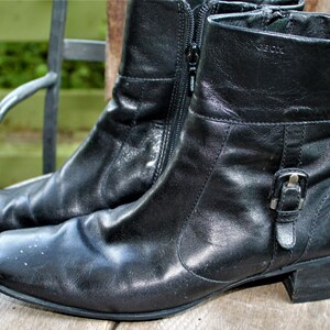 May include: A pair of black leather ankle boots with a buckle detail on the side. The boots have a low heel and a zipper on the inside of each boot.