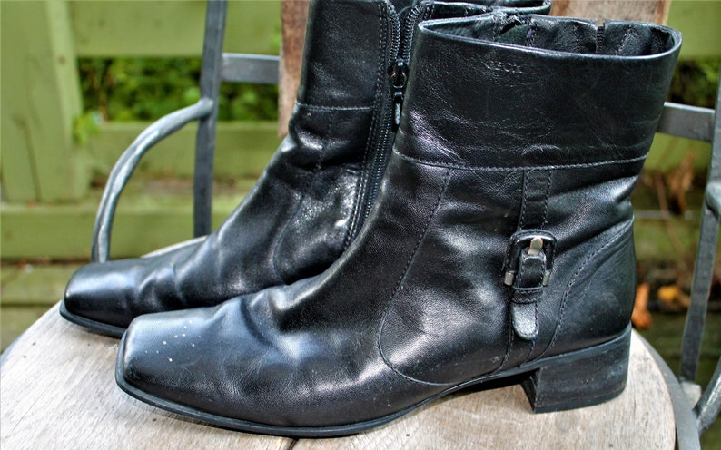May include: A pair of black leather ankle boots with a buckle detail on the side. The boots have a low heel and a zipper on the inside of each boot.
