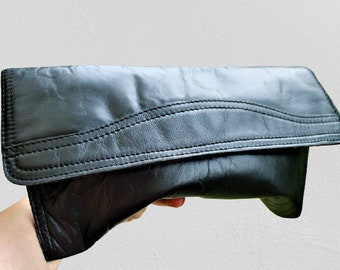Könnte beinhalten: Eine schwarze Leder-Clutch mit Klappenverschluss und dekorativen Nähten. Die Clutch ist in der Mitte gefaltet und wird in einer Hand gehalten.