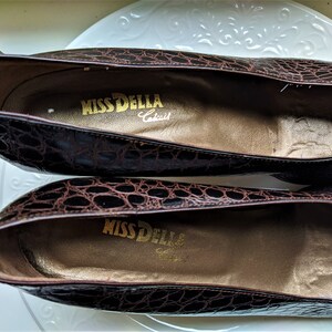 May include: A pair of brown crocodile-embossed leather shoes with a gold label that reads "Miss Della,  Crafted".