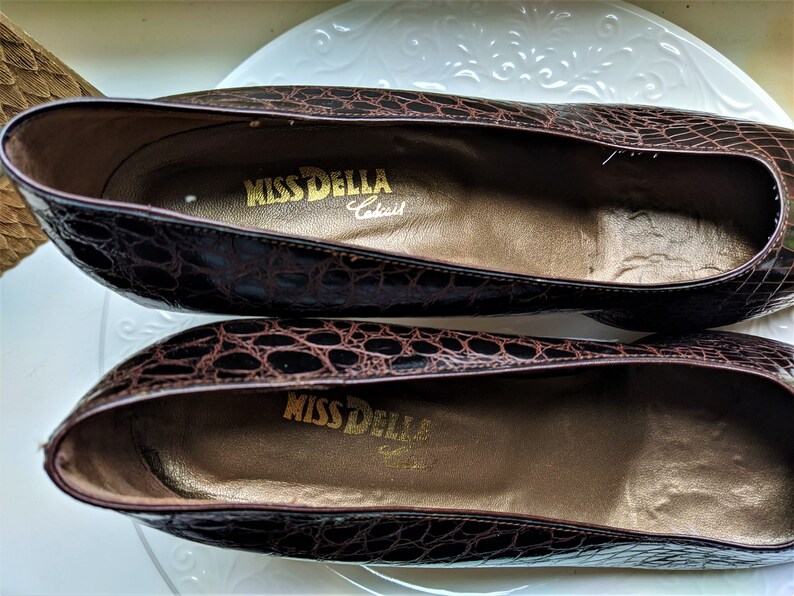 May include: A pair of brown crocodile-embossed leather shoes with a gold label that reads "Miss Della,  Crafted".