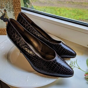 May include: A pair of brown crocodile-embossed leather pumps with pointed toes and stiletto heels. The shoes are on a white plate.