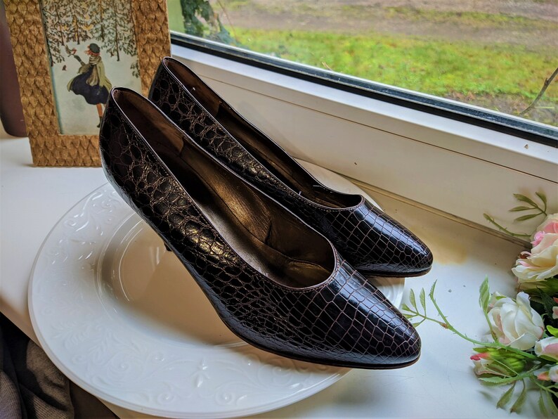 May include: A pair of brown crocodile-embossed leather pumps with pointed toes and stiletto heels. The shoes are on a white plate.